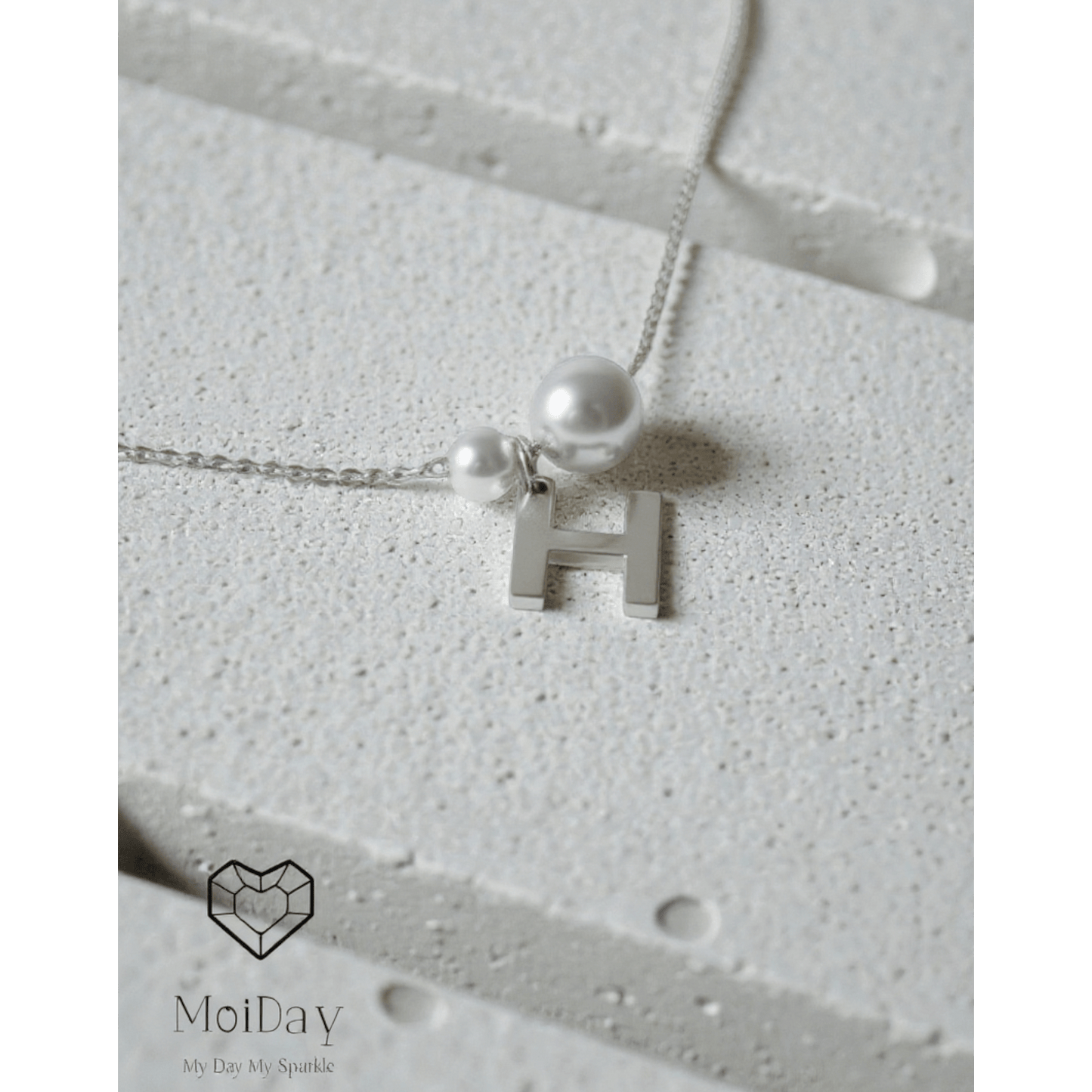 Bracelet with a letter 'H' pendant and pearl on a textured surface, branded 'MoiDay'.