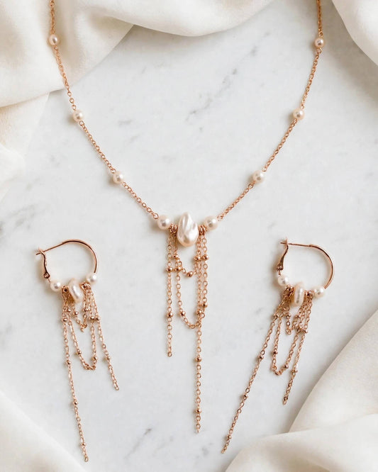 Rose gold jewelry set featuring a necklace, matching hoop earrings, and dangling chain earrings, styled on a white marble surface with draped white fabric.