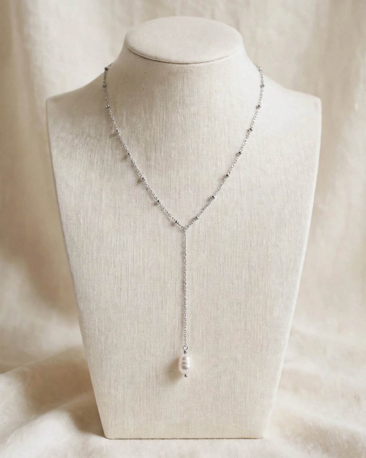 A delicate silver Y-necklace with small bead accents and a single white pearl pendant, displayed on a cream fabric-covered jewelry bust against a soft beige backdrop.