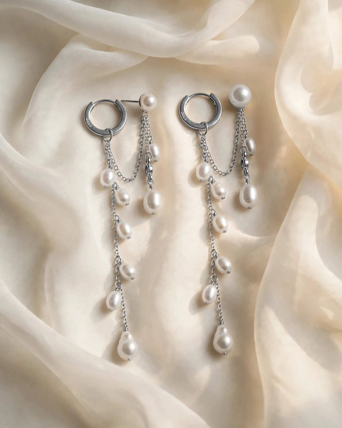 A pair of silver hoop dangle earrings with multiple white pearl drops, arranged on draped cream-colored fabric.
