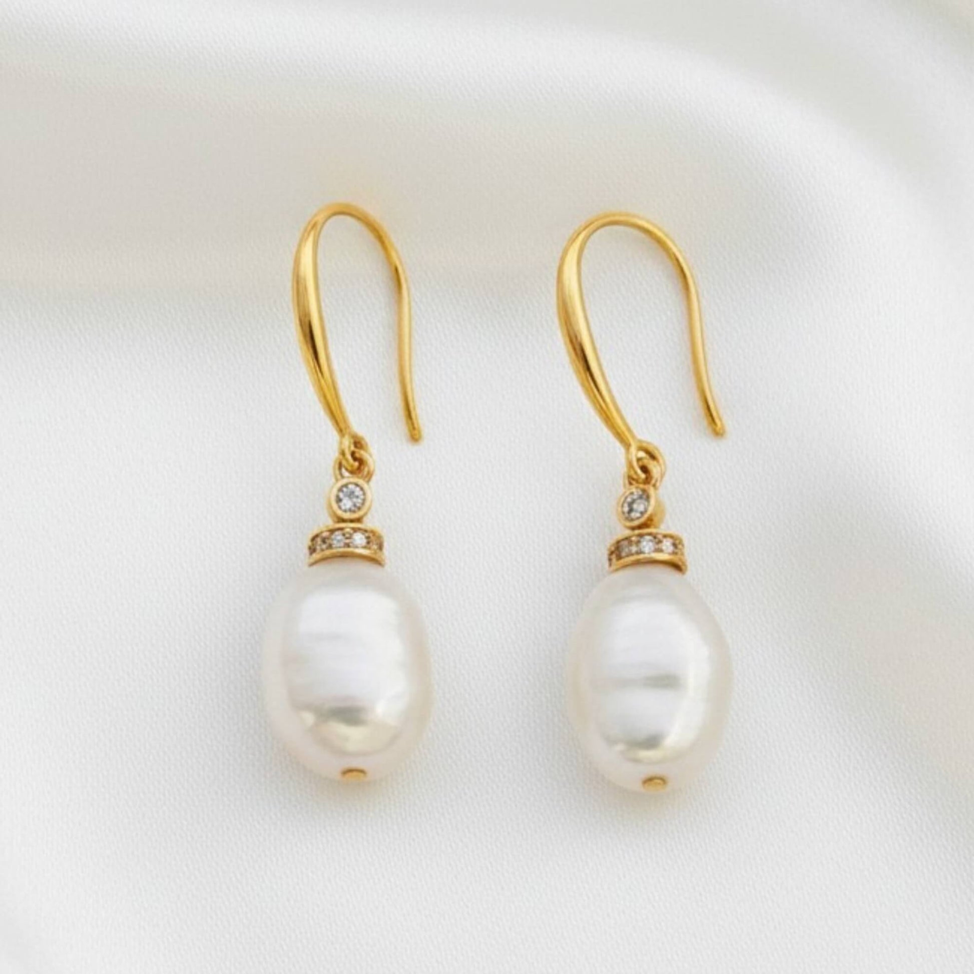Gold hook earrings with white pearl and zirconia crystal photographed on soft white fabric, quiet luxury style.