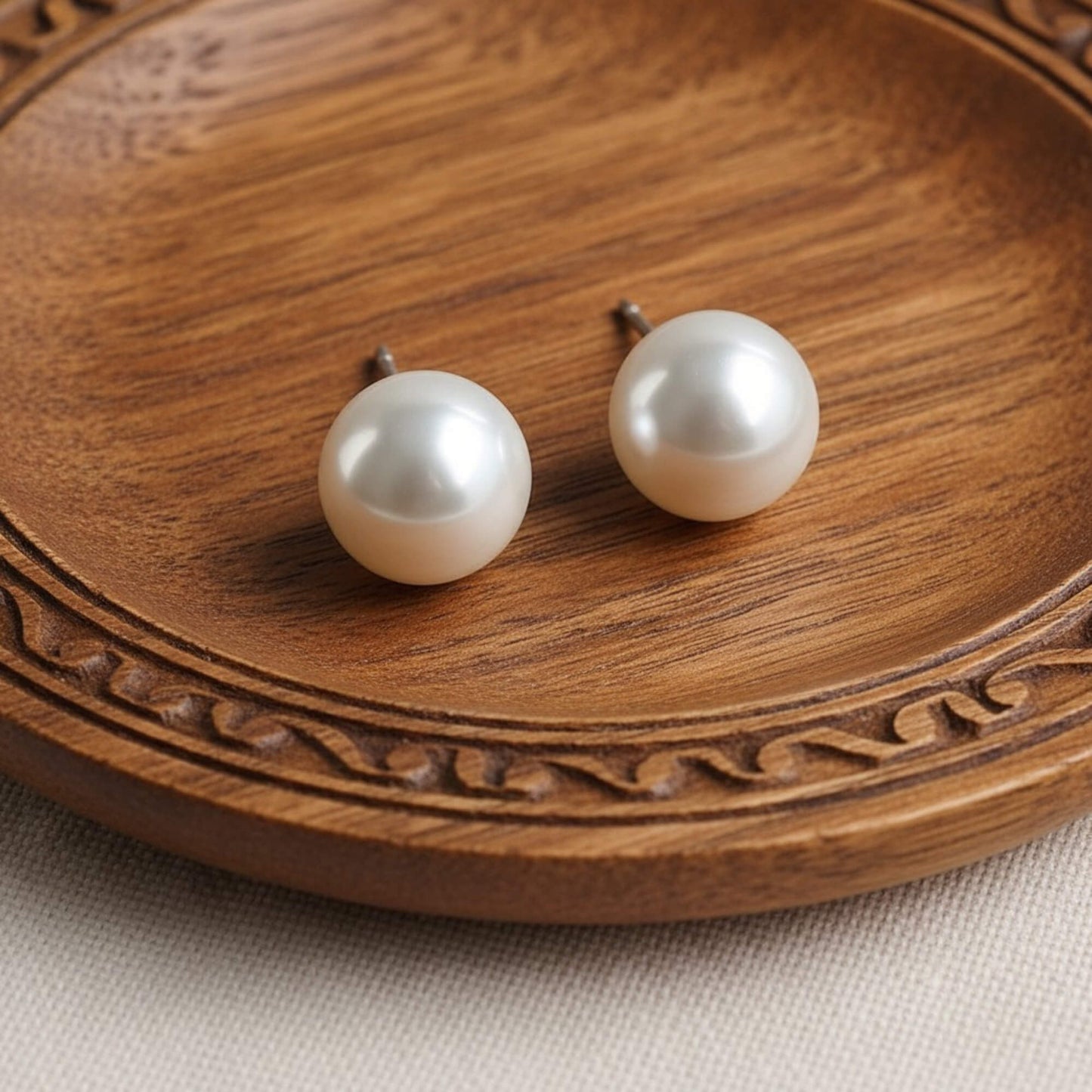  A pair of classic white pearl stud earrings placed on beige textured fabric with soft natural lighting.