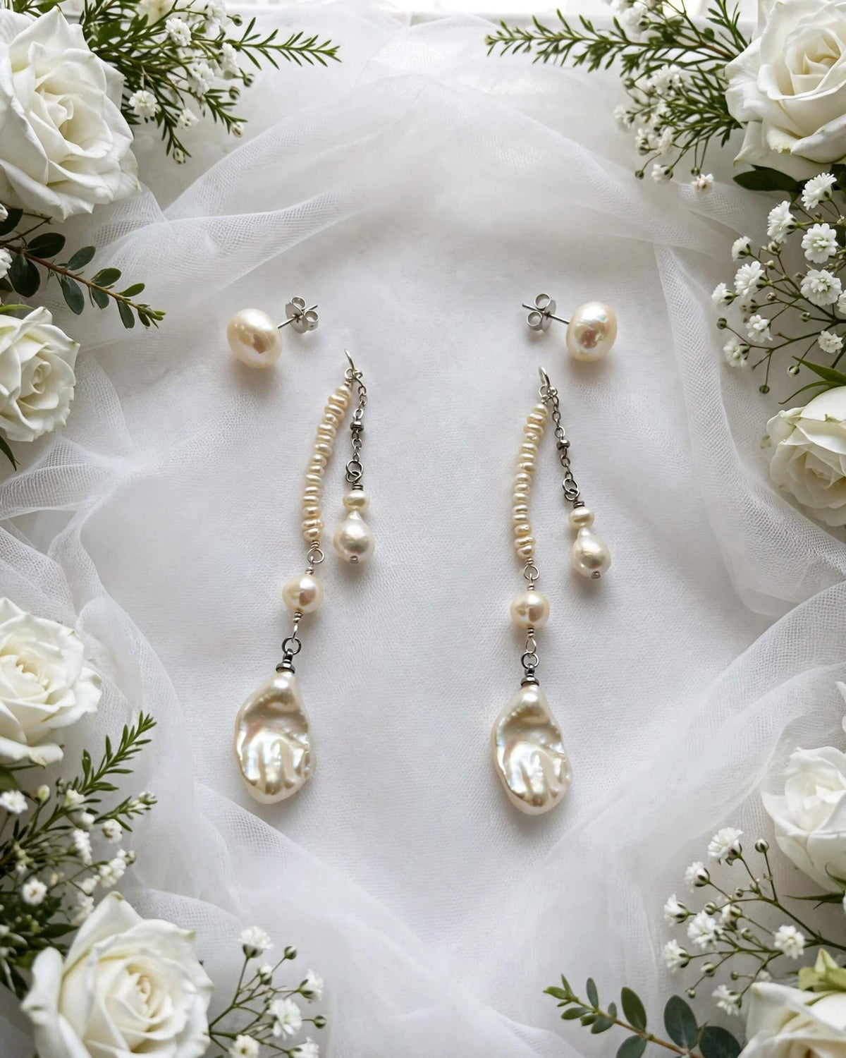 A pair of long dangle earrings and matching stud earrings made from cream-colored baroque pearls and small silver-toned chain links, arranged on white tulle fabric and surrounded by white roses and baby's breath.