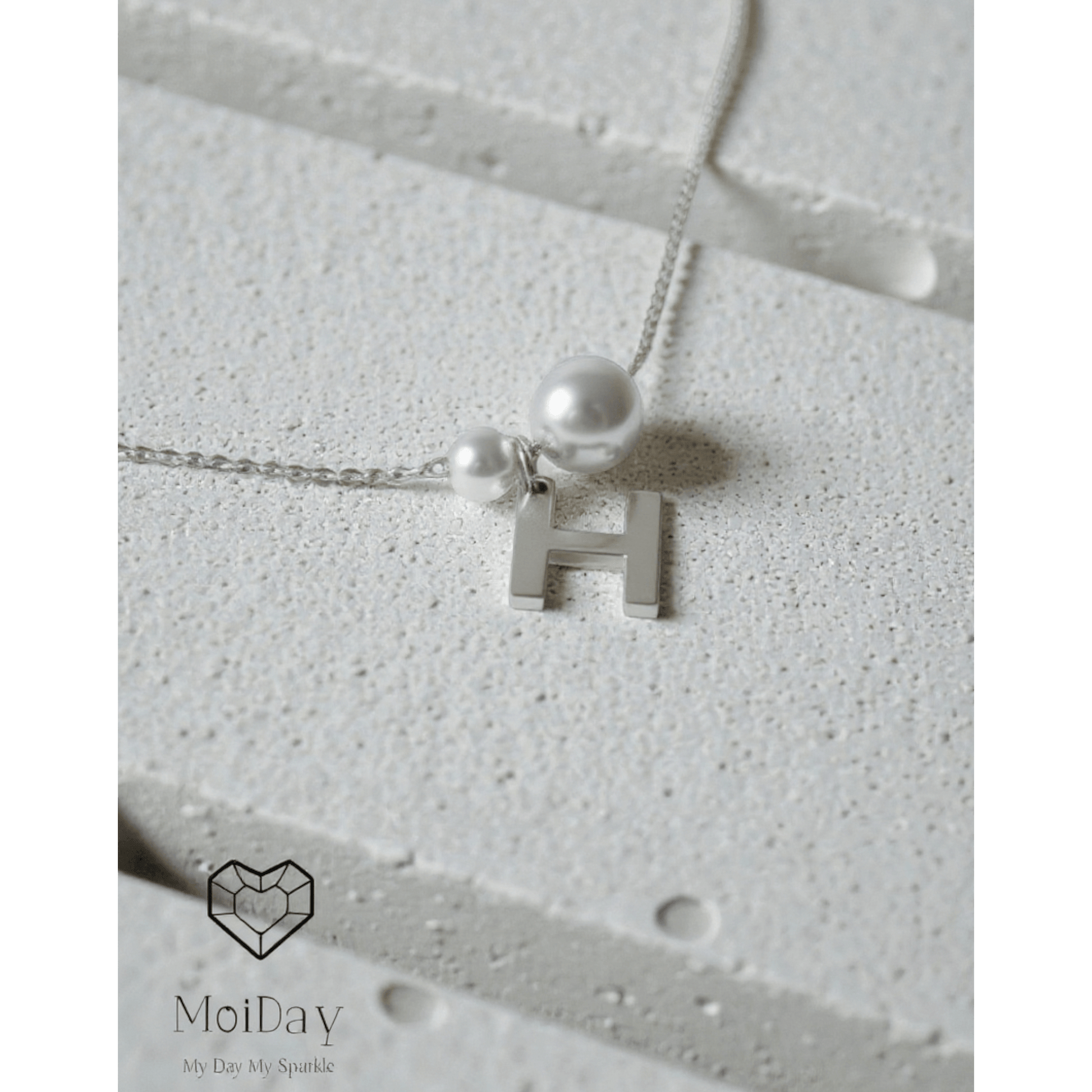 Bracelet with a letter 'H' pendant and pearl on a textured surface, branded 'MoiDay'.