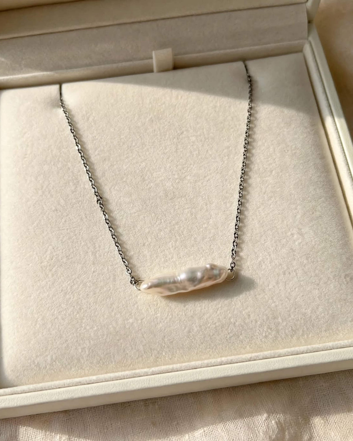 A silver chain necklace with a single irregular baroque pearl pendant, displayed inside an open cream-colored jewelry box.
