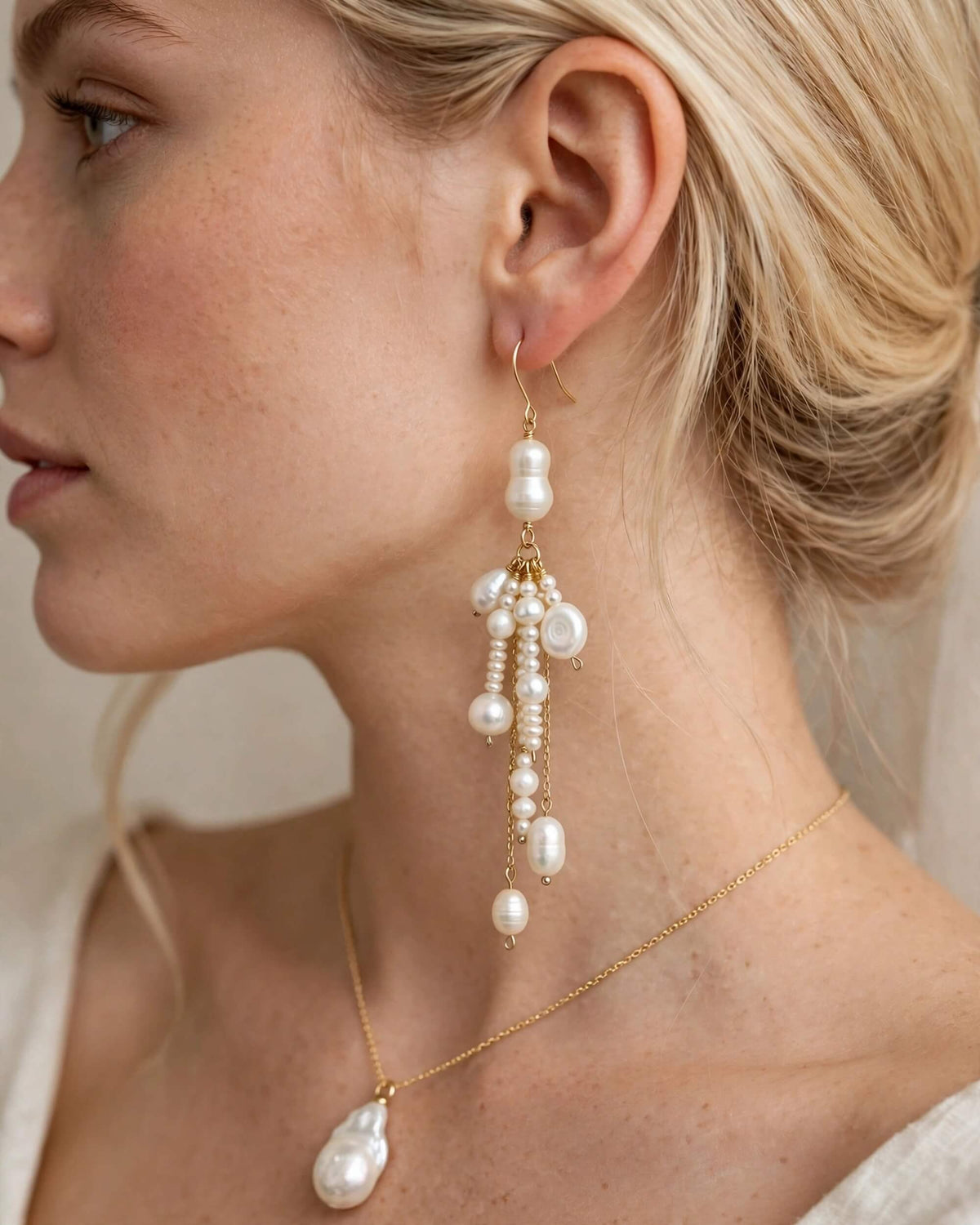 A close-up side profile of a blonde woman wearing a long gold drop earring with multiple white baroque pearls, along with a matching gold necklace with a teardrop pearl pendant.
