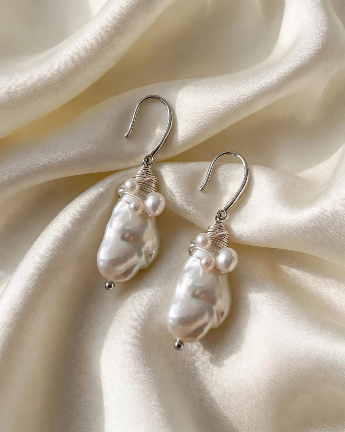 A pair of silver dangle earrings with irregular white baroque pearls and small pearl-like beads, displayed on draped cream satin fabric.