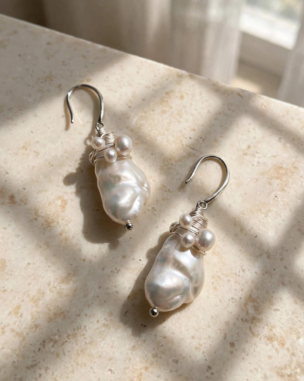 A pair of silver drop earrings featuring irregular white baroque pearls and small wrapped pearl accents, displayed on a textured beige stone surface with soft window light and shadow patterns.