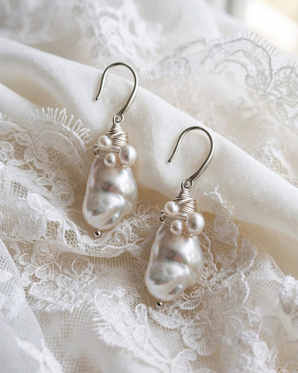 A pair of silver drop earrings with irregular white baroque pearls and small pearl-like beads, displayed on white lace fabric.