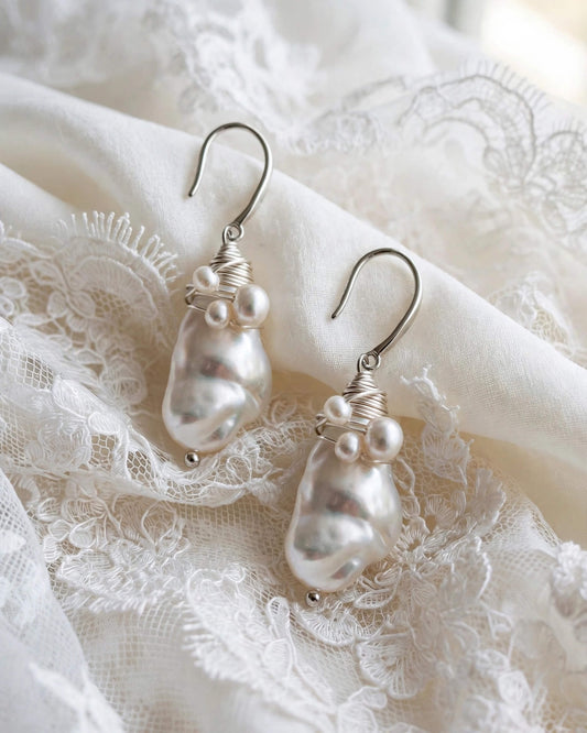 A pair of silver drop earrings with irregular white baroque pearls and small pearl-like beads, displayed on white lace fabric.