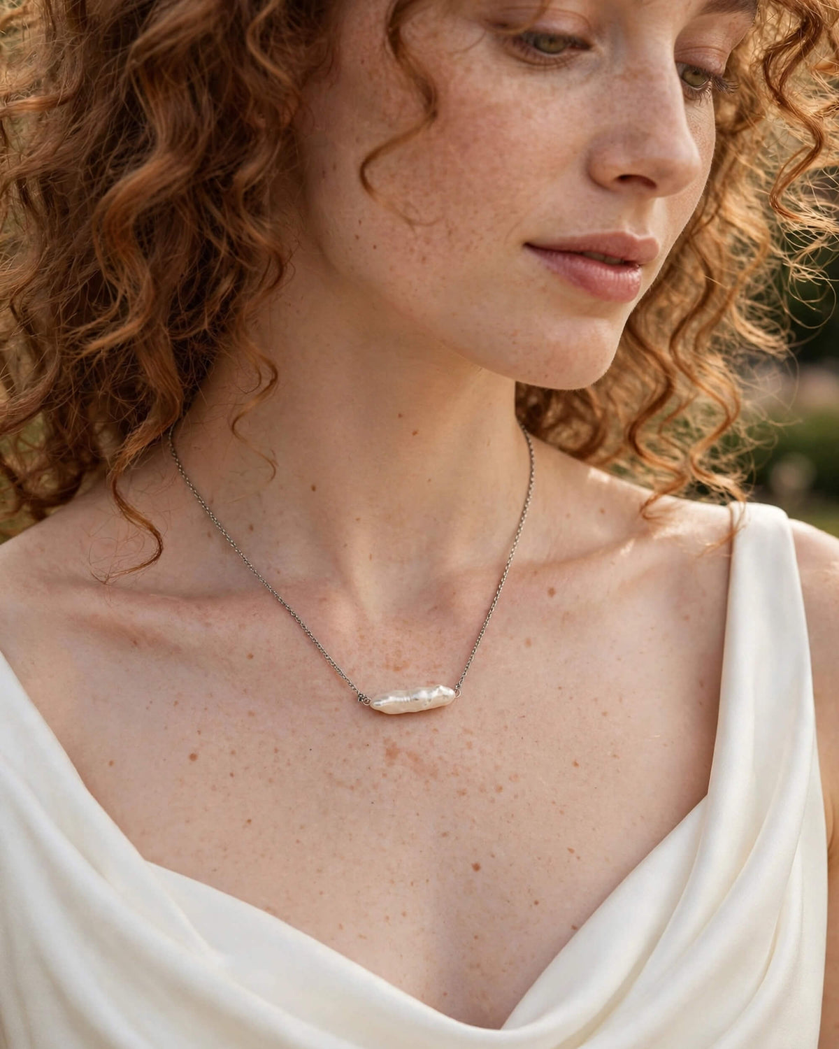 A close-up lifestyle shot of a woman with curly red hair wearing a silver chain necklace with a natural baroque pearl pendant, paired with a white draped top.