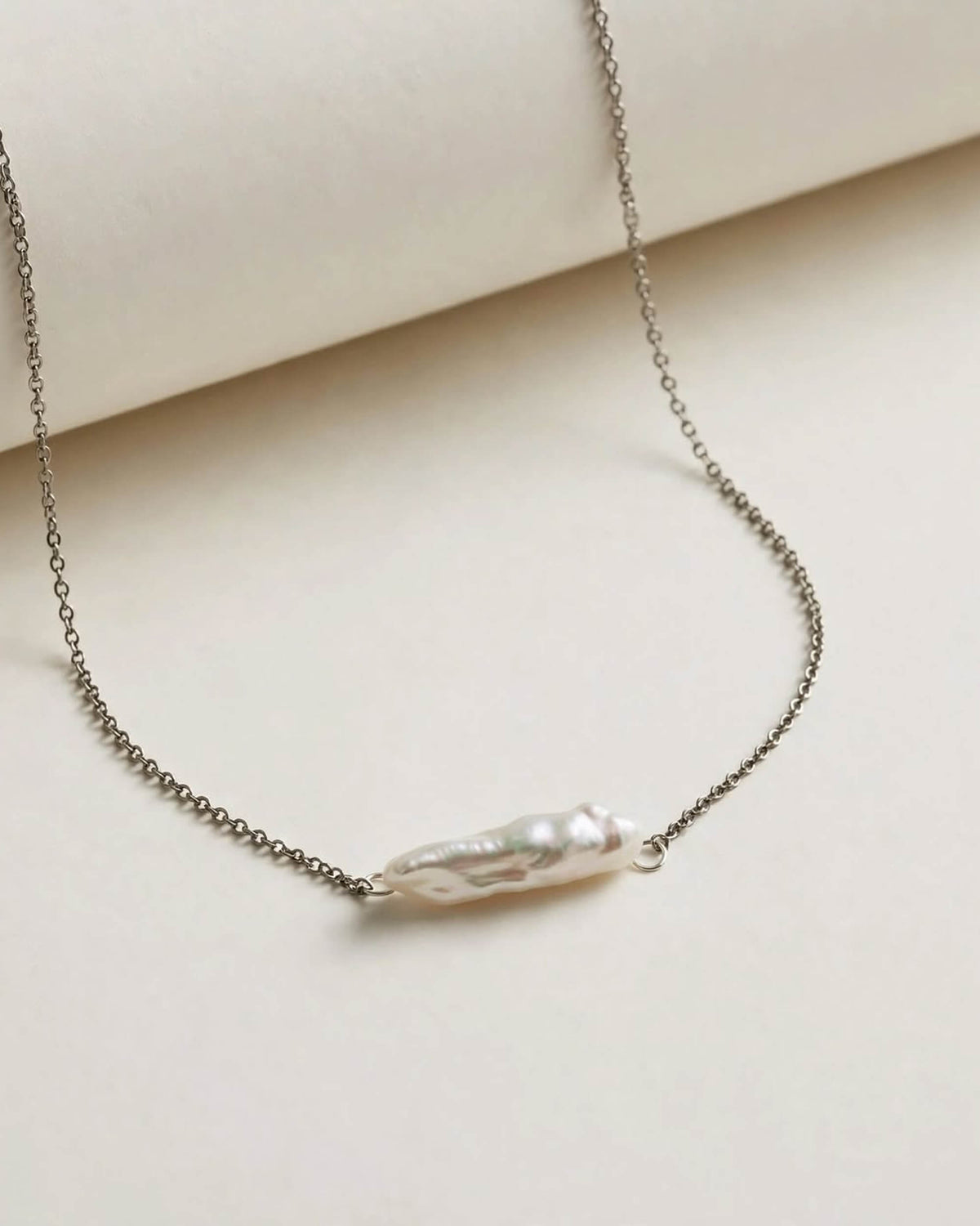 A delicate silver chain necklace featuring a single irregular, baroque-style white pearl pendant, displayed on a clean, light beige surface.