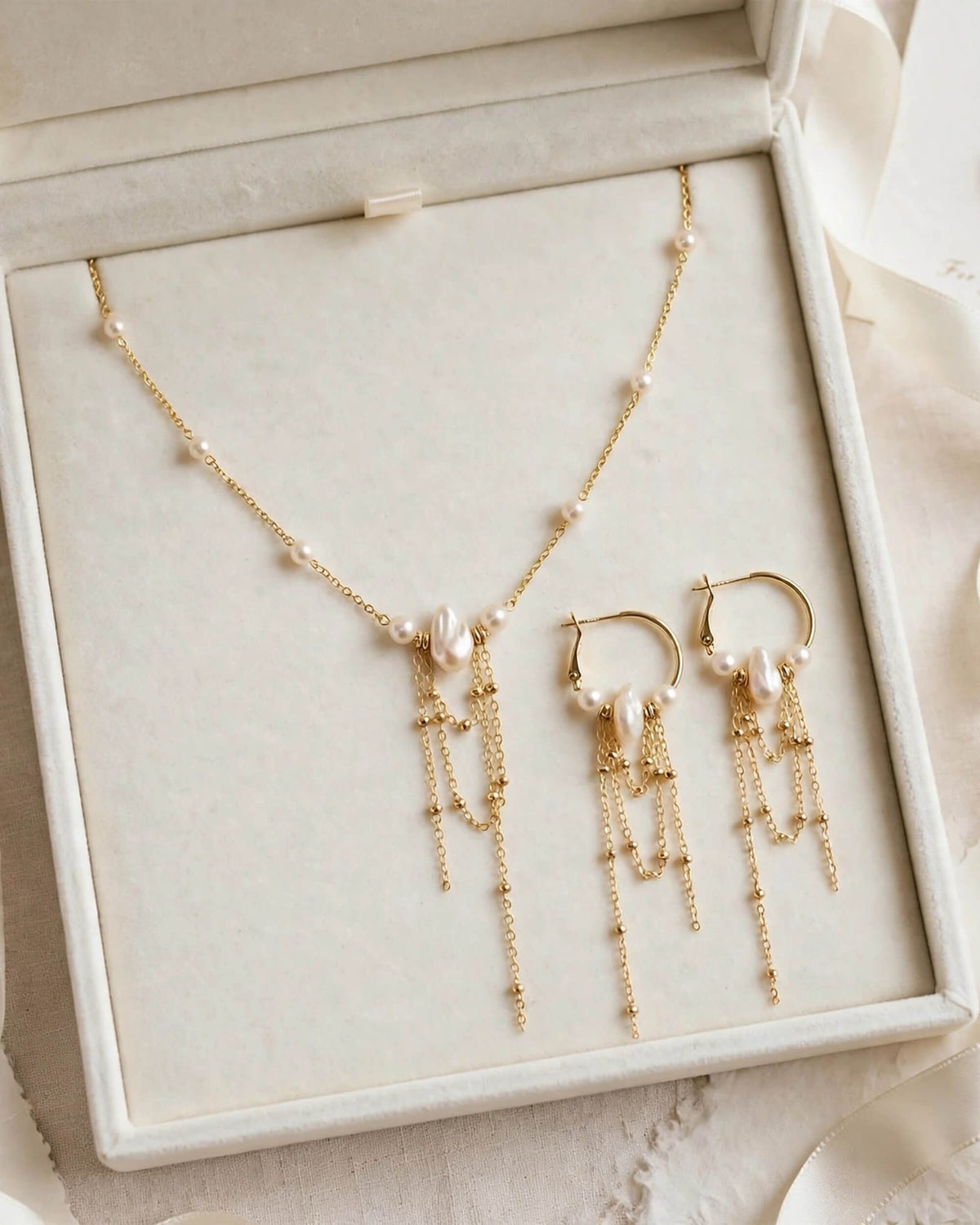 Gold jewelry set with a necklace and matching earrings displayed in an open cream-colored presentation box. The set features delicate gold chains, pearl-like beads, and dangling chain and pearl accents.