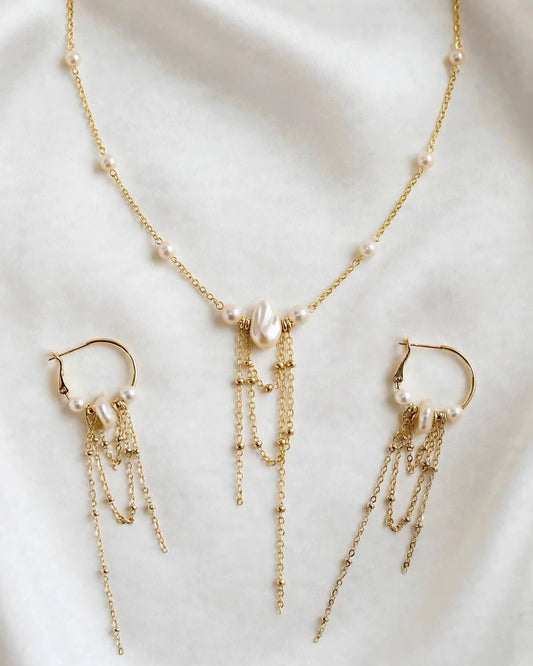 Gold-tone jewelry set featuring a delicate necklace and matching drop earrings, both adorned with small white pearls and layered chain drapes, displayed on a soft white fabric background.