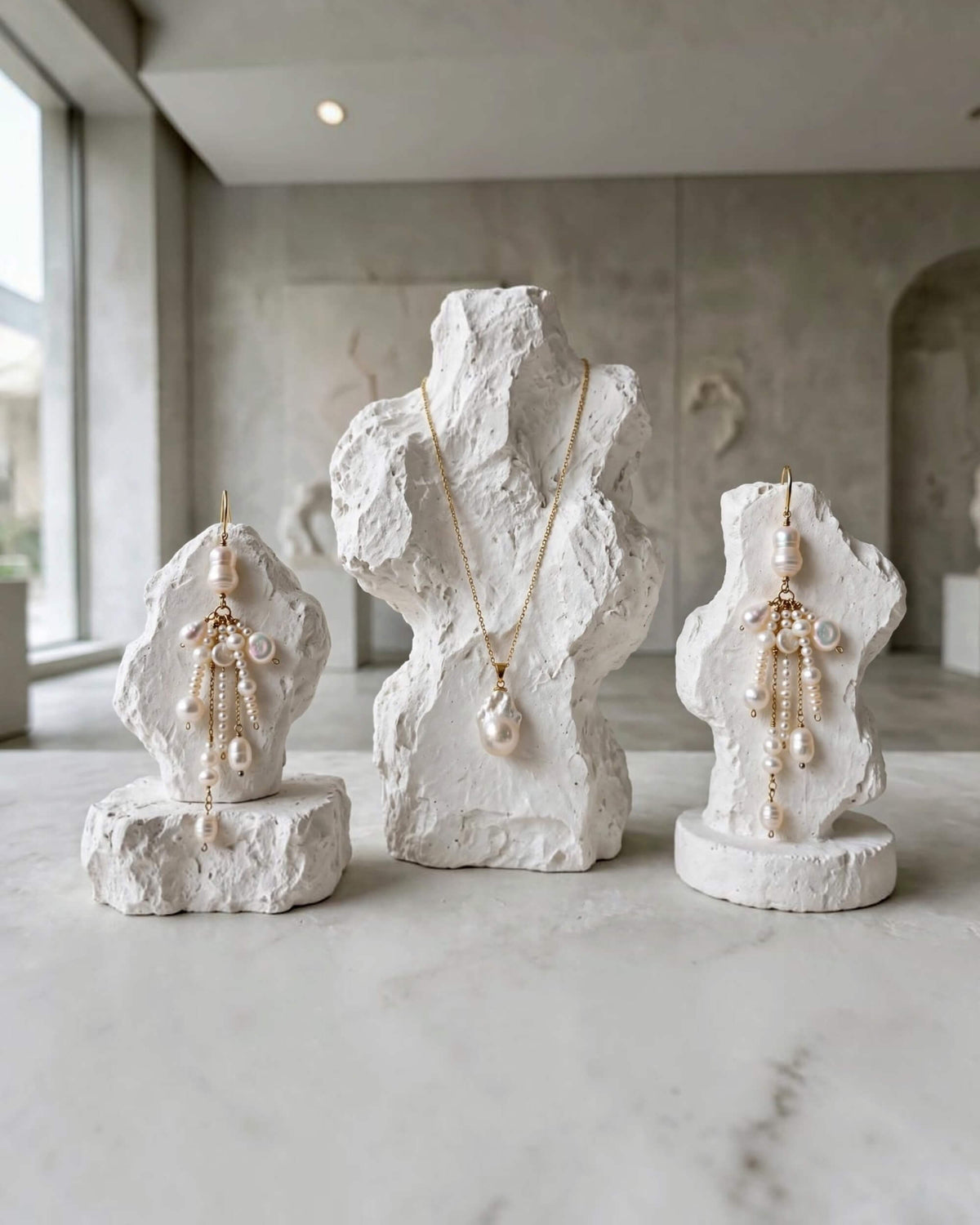 Three white stone bust displays showcasing gold jewelry: a necklace with a large pearl pendant in the center, and matching pearl drop earrings on both sides, set on a light marble surface in a minimalist, modern interior.