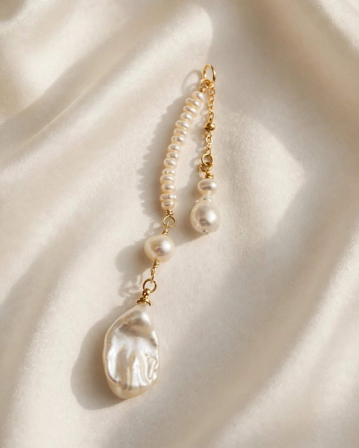 A gold-tone beaded chain accessory with multiple white baroque pearls and a large irregular pearl dangle, laid on draped cream satin fabric.