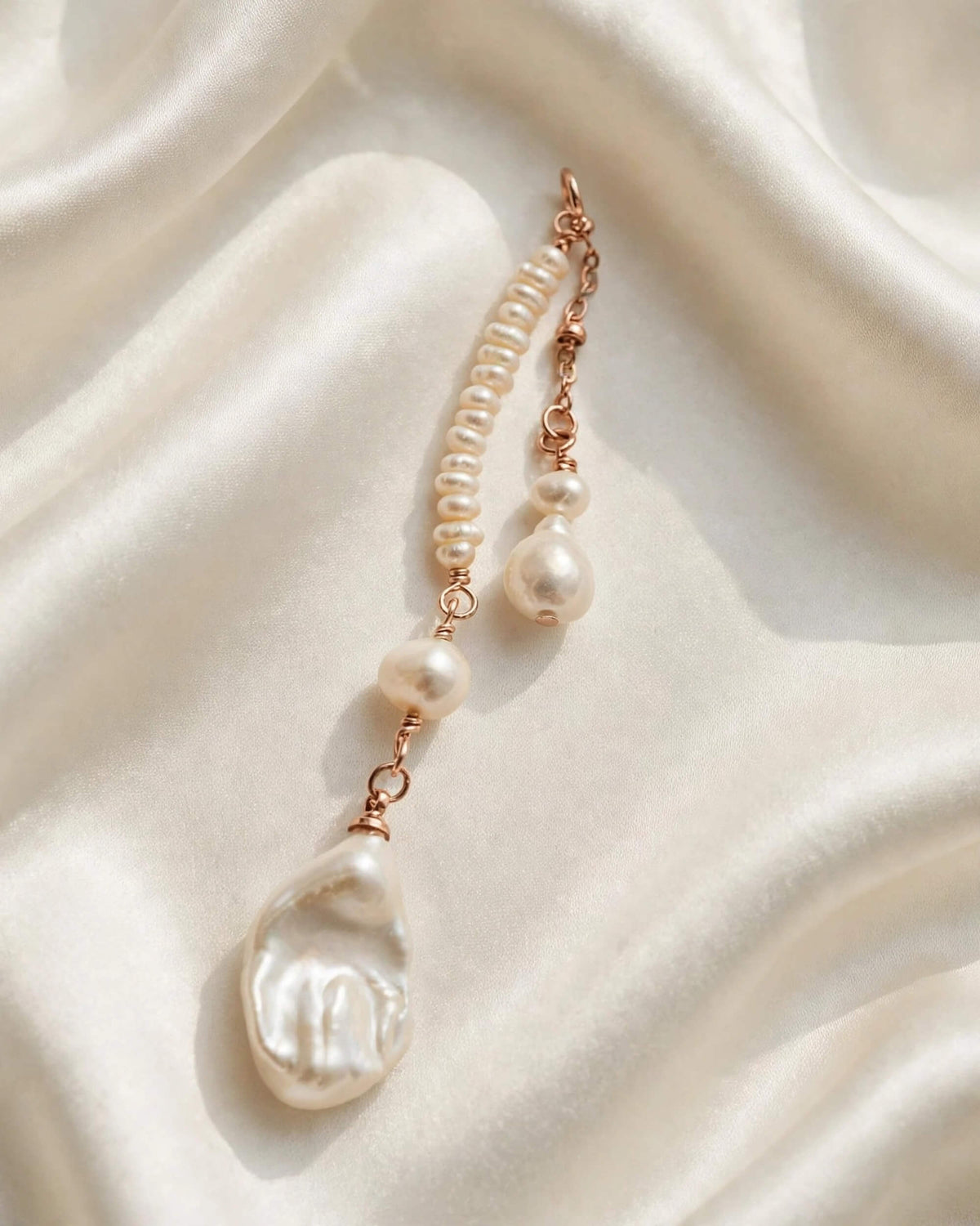 A baroque pearl drop earring with a rose gold chain and spacer beads, displayed on a draped cream satin fabric.