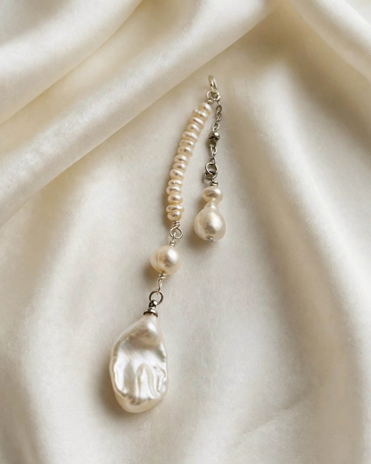 A single pearl drop earring with a beaded chain, a small pearl bead, and a large baroque-style pearl drop, displayed on draped cream satin fabric.