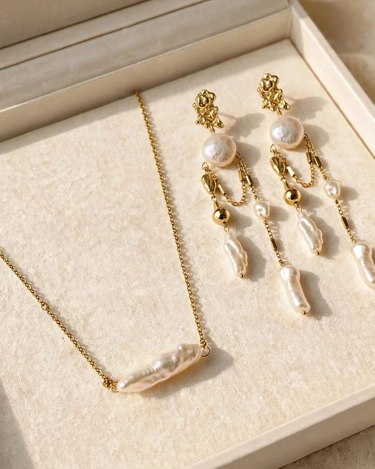 Gold jewelry set with a baroque pearl necklace and matching drop earrings, displayed on a cream-colored jewelry box.
