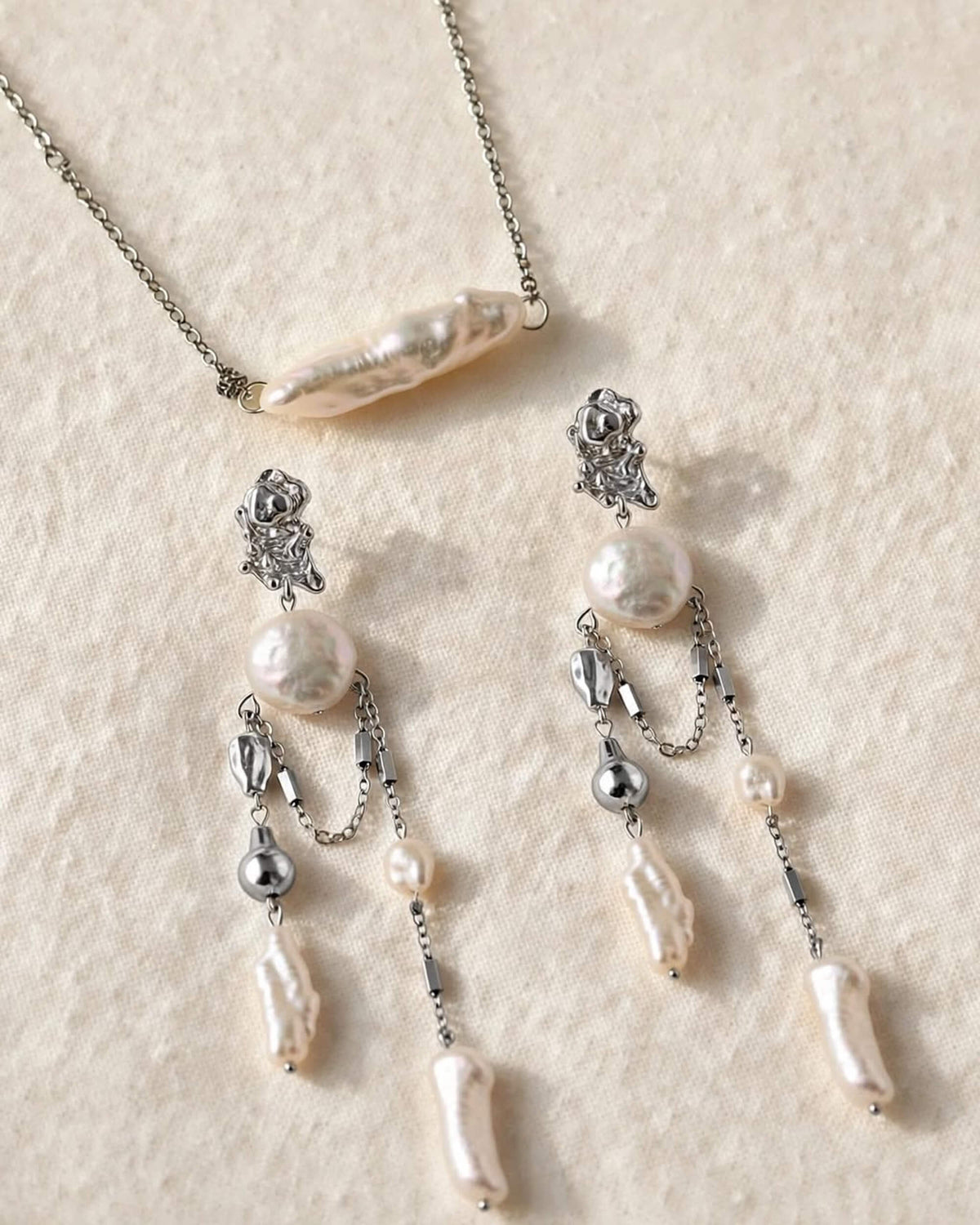 A silver-toned jewelry set featuring a necklace and matching drop earrings with irregular white baroque pearls and small metallic beads, arranged on a textured cream fabric background.