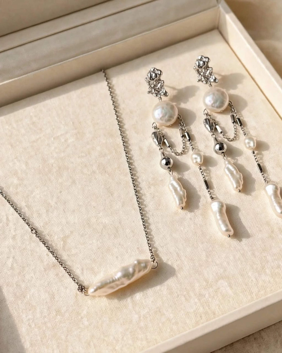 A jewelry set featuring a silver chain necklace with a baroque pearl pendant and matching silver drop earrings with baroque pearls, displayed on a beige textured jewelry box.