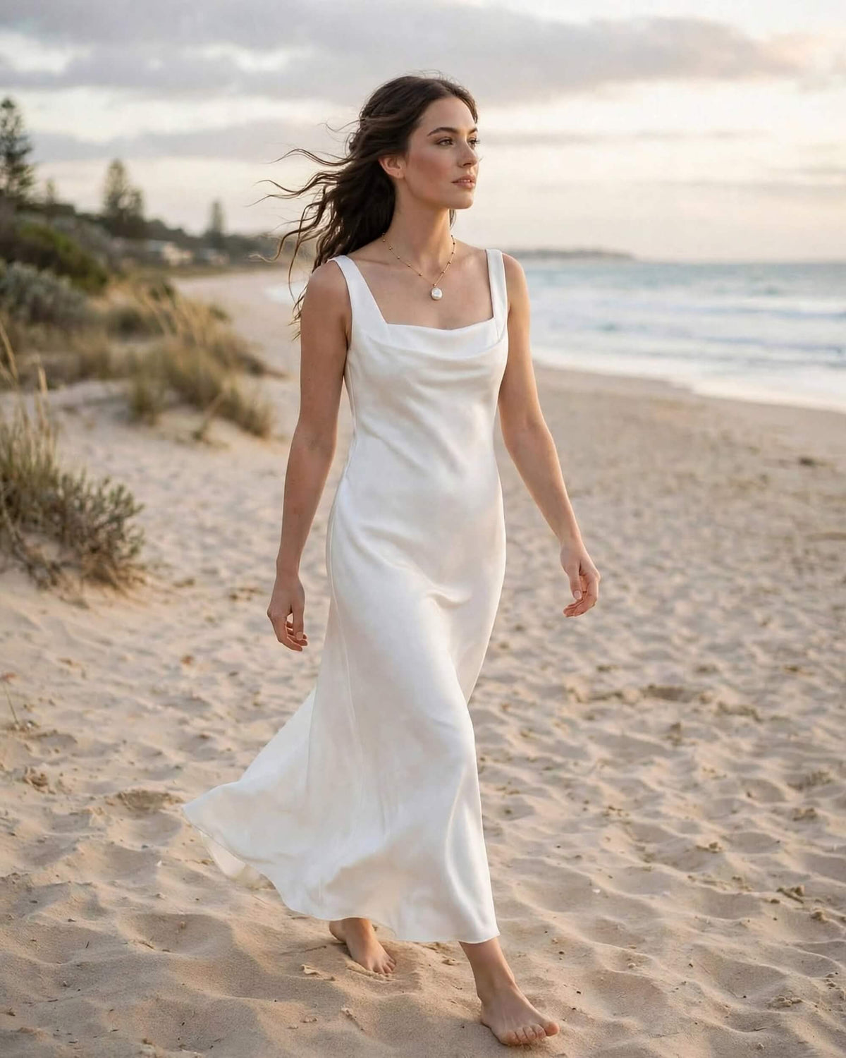 A woman walking barefoot on a sandy beach wearing a flowing white maxi slip dress with a draped neckline.