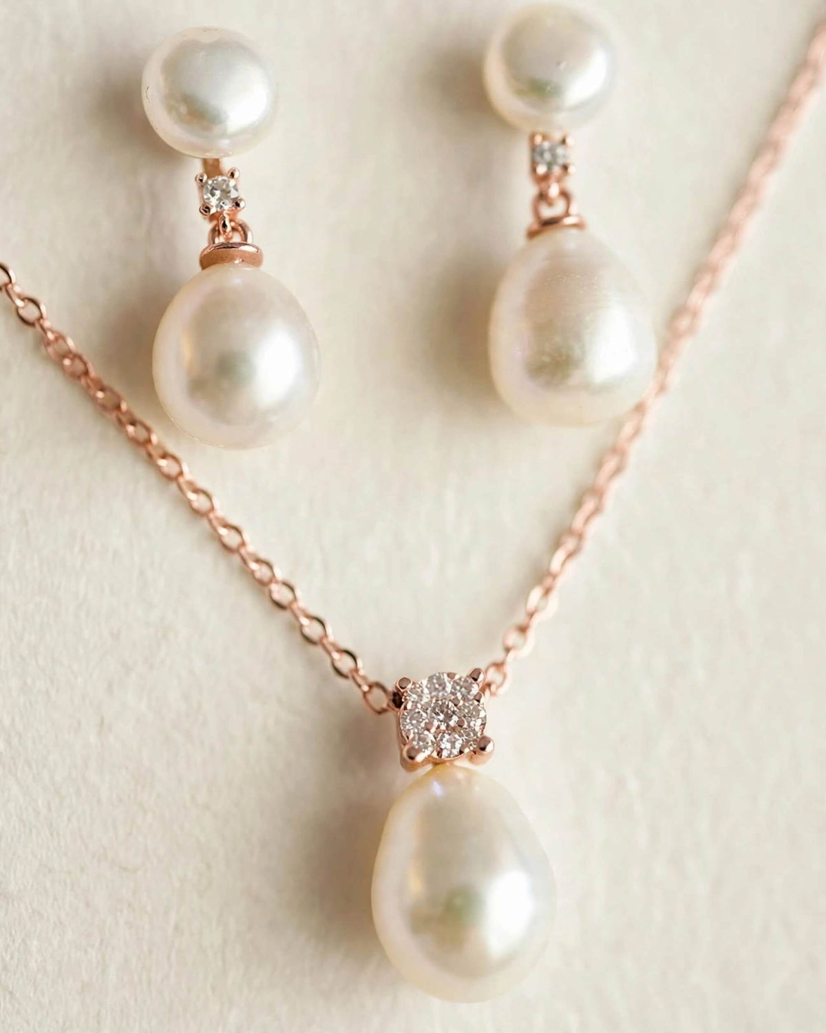 Rose gold jewelry set featuring a necklace and matching drop earrings with large white freshwater pearls and small clear crystal accents, displayed on a textured cream background.