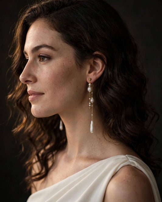 A woman in profile wearing a long pearl drop earring with a gold chain and teardrop pendant, photographed against a dark studio background.