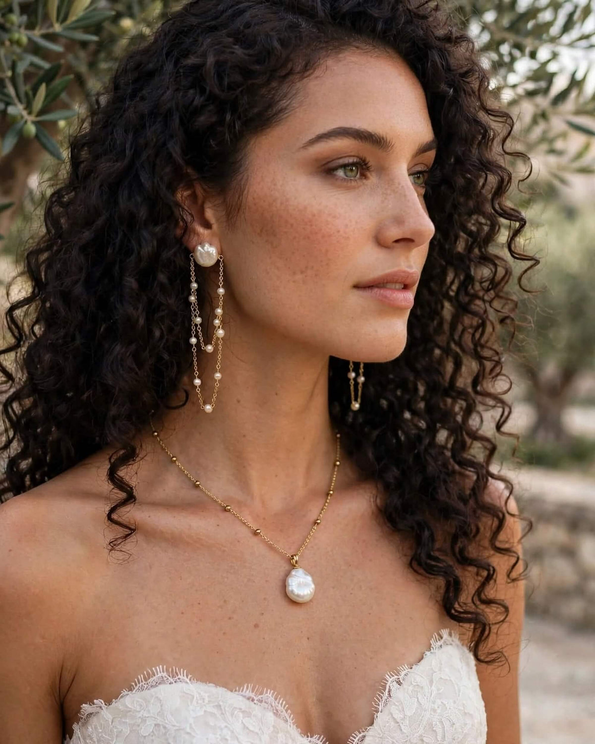 A woman with long, curly dark hair is shown in profile wearing a gold necklace with a large white pearl pendant and matching pearl drop earrings. She is wearing a white lace strapless top, with a blurred outdoor background featuring green foliage.