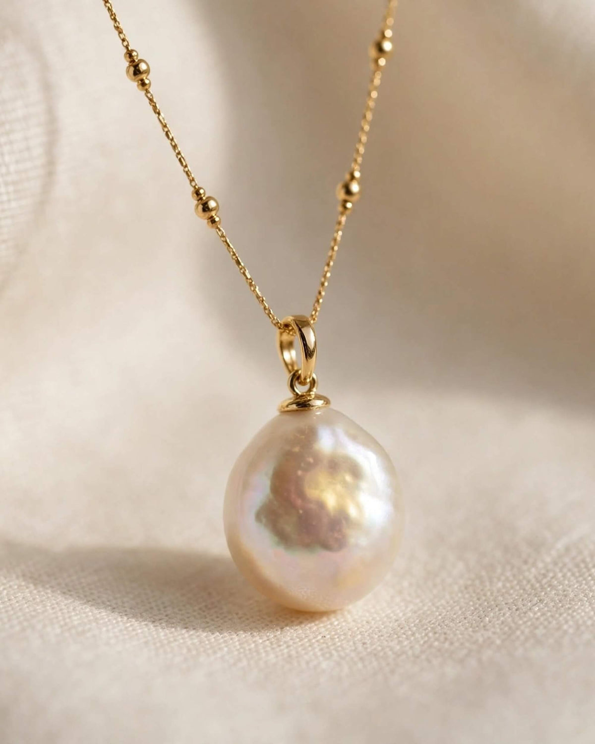 A gold chain necklace with a single irregular, lustrous white pearl pendant, displayed on a soft beige fabric background.
