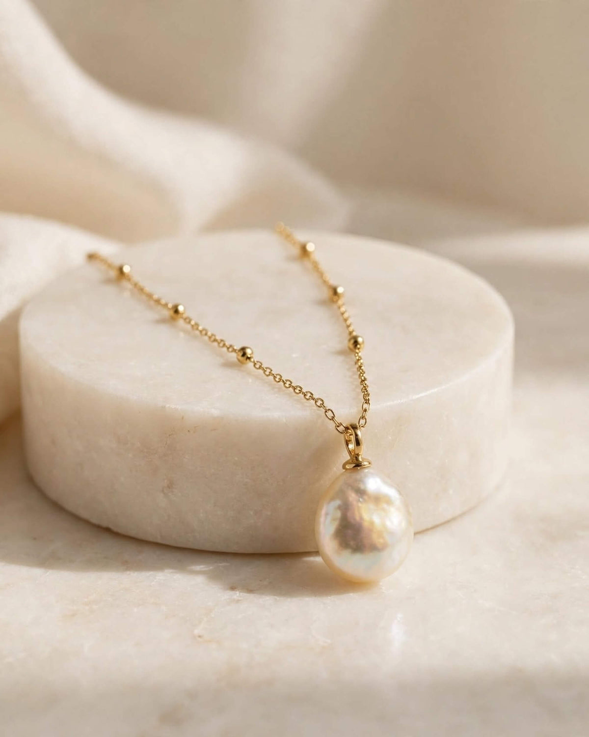 A delicate gold chain necklace with a large, teardrop-shaped baroque pearl pendant, displayed on a round white marble pedestal.