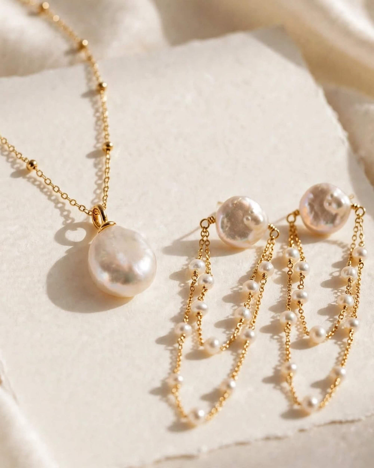 Gold-tone jewelry set featuring a necklace and matching earrings with irregular white freshwater pearls, styled on a textured cream paper background.
