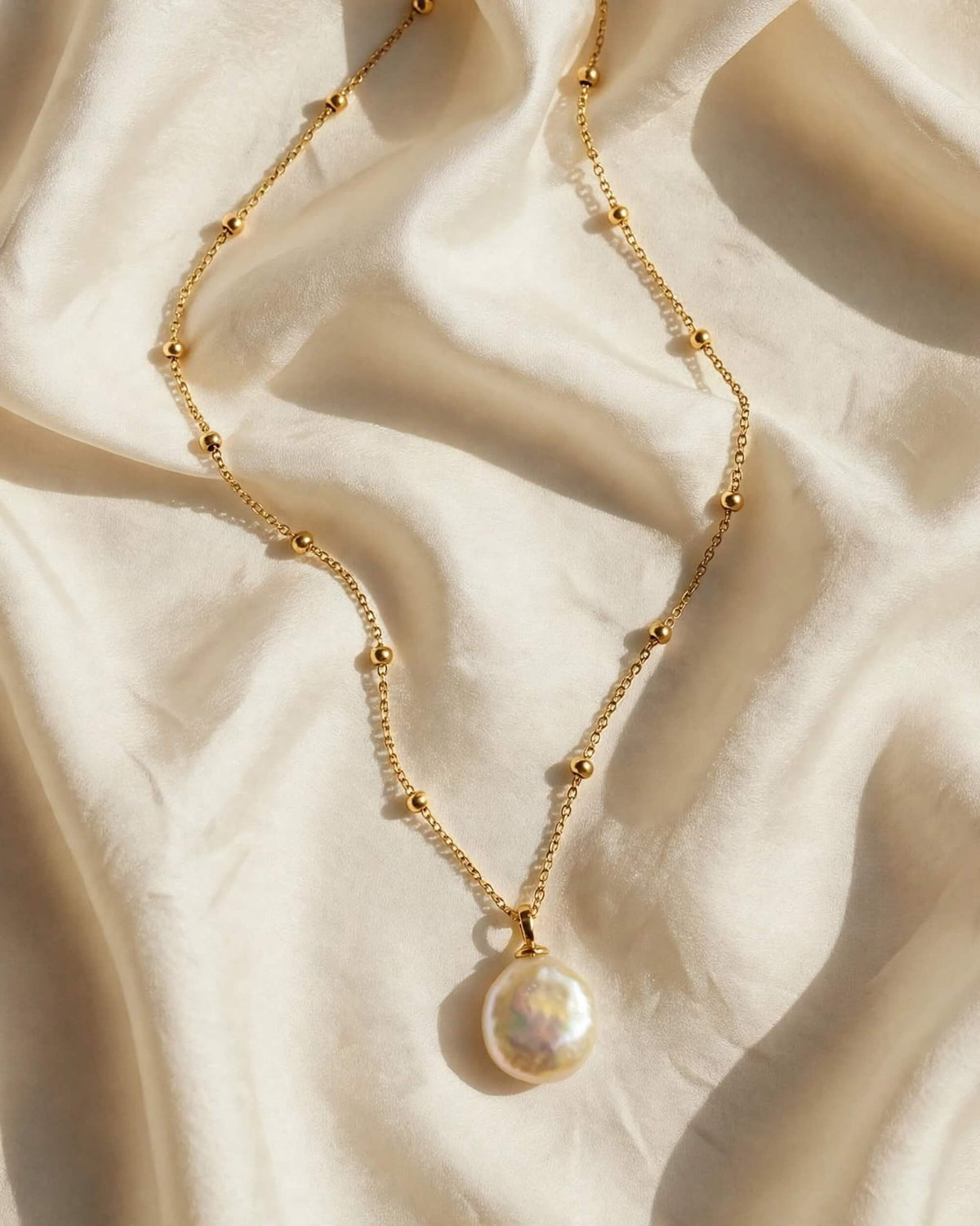 A delicate gold chain necklace with small gold beads and a large, lustrous white pearl pendant, draped elegantly on draped cream satin fabric.