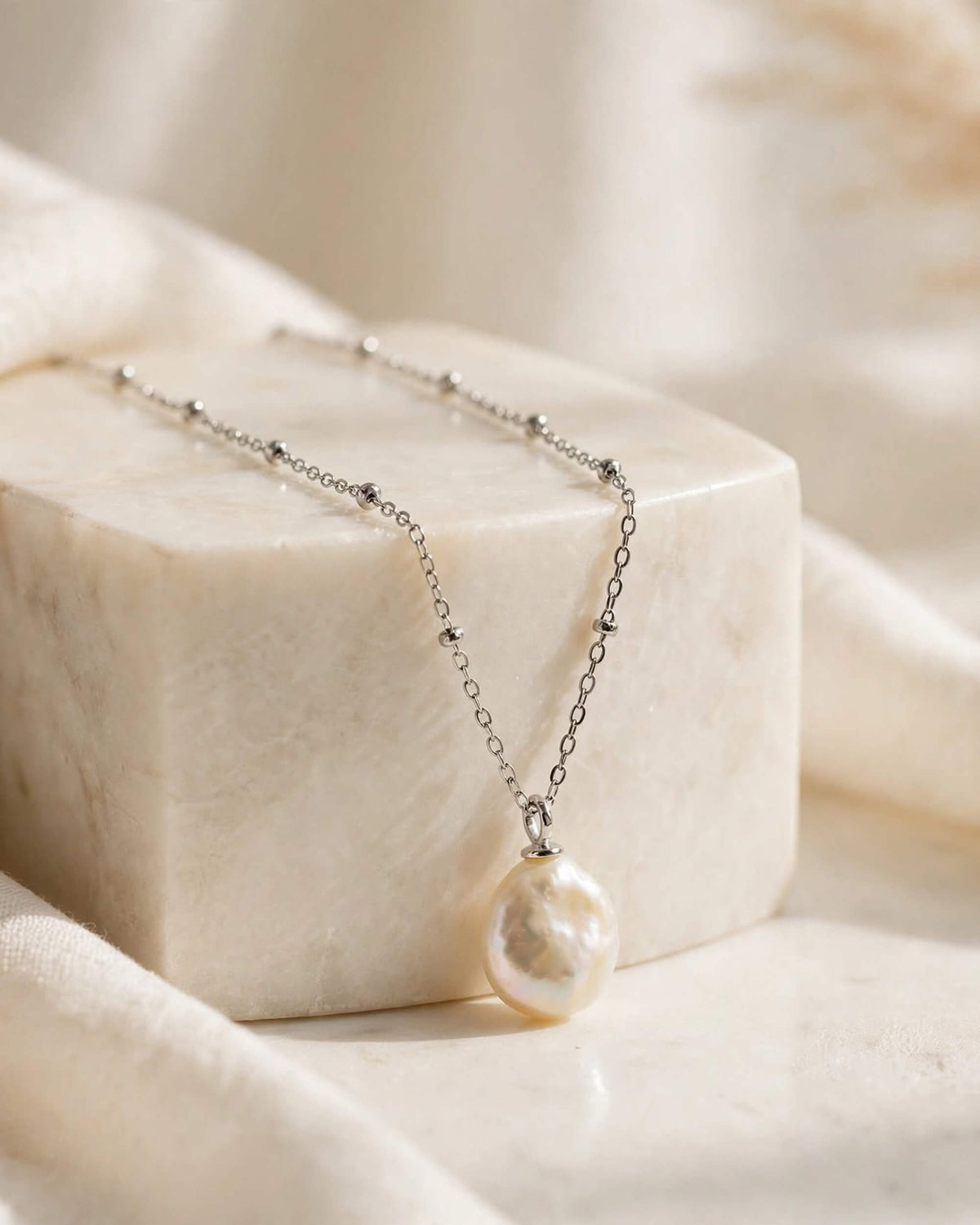 A delicate silver chain necklace with a large, irregular white pearl pendant, displayed on a cream-colored marble block with soft draped fabric in the background.