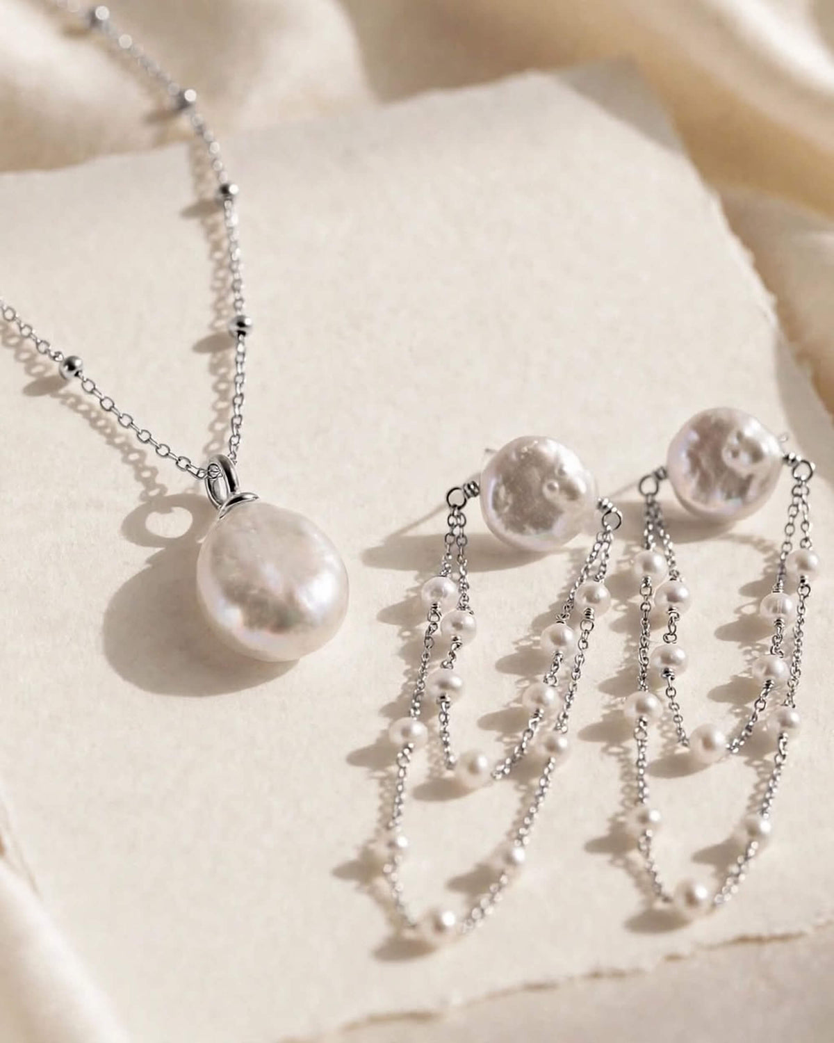 Silver-toned jewelry set featuring a necklace and matching earrings with irregular white pearls and layered bead chains, arranged on a textured cream surface with soft fabric in the background.