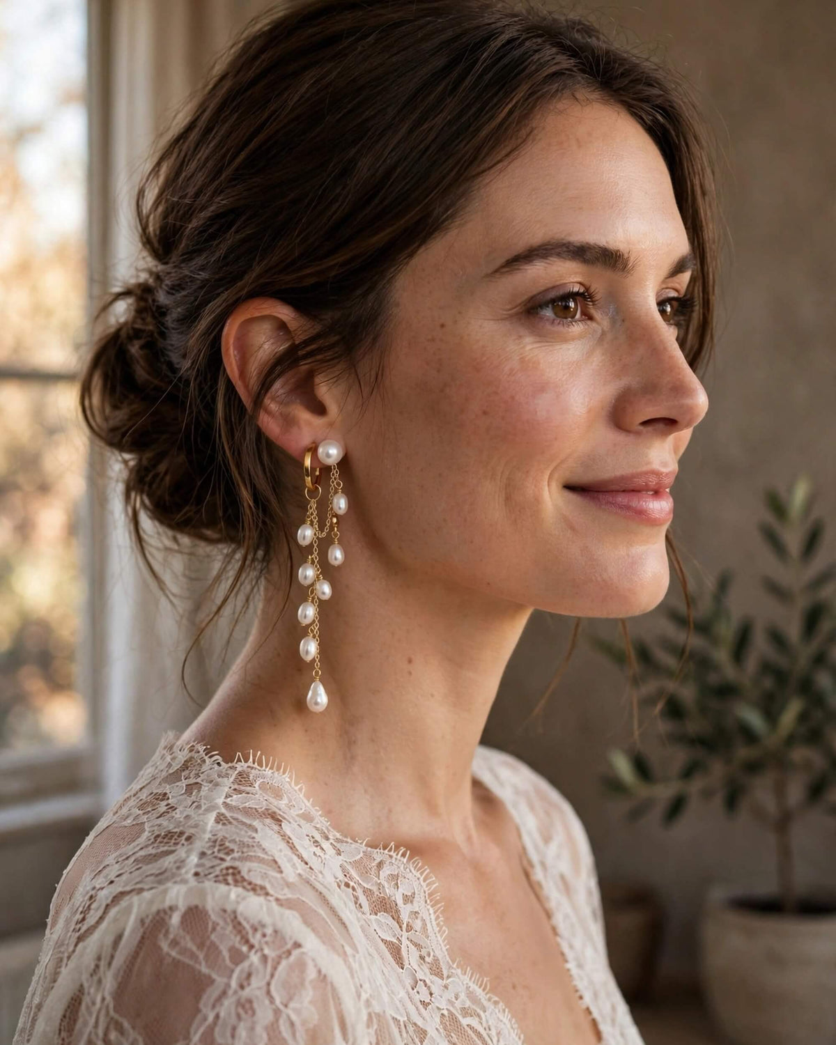 A woman in profile wearing long gold drop earrings with a series of white pearl-like beads, photographed indoors near a window with soft natural light.
