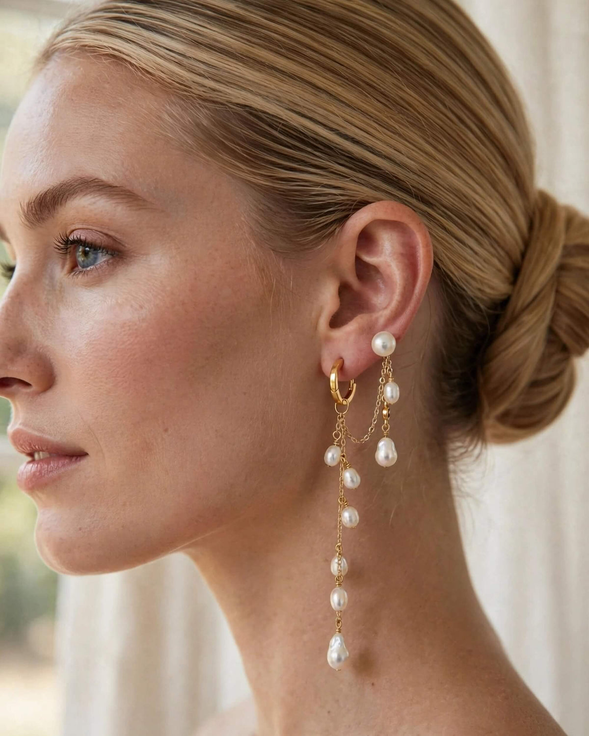 A close-up side profile of a woman with blonde hair in a bun, wearing a pair of gold hoop earrings with dangling pearl-like beads and chain details.