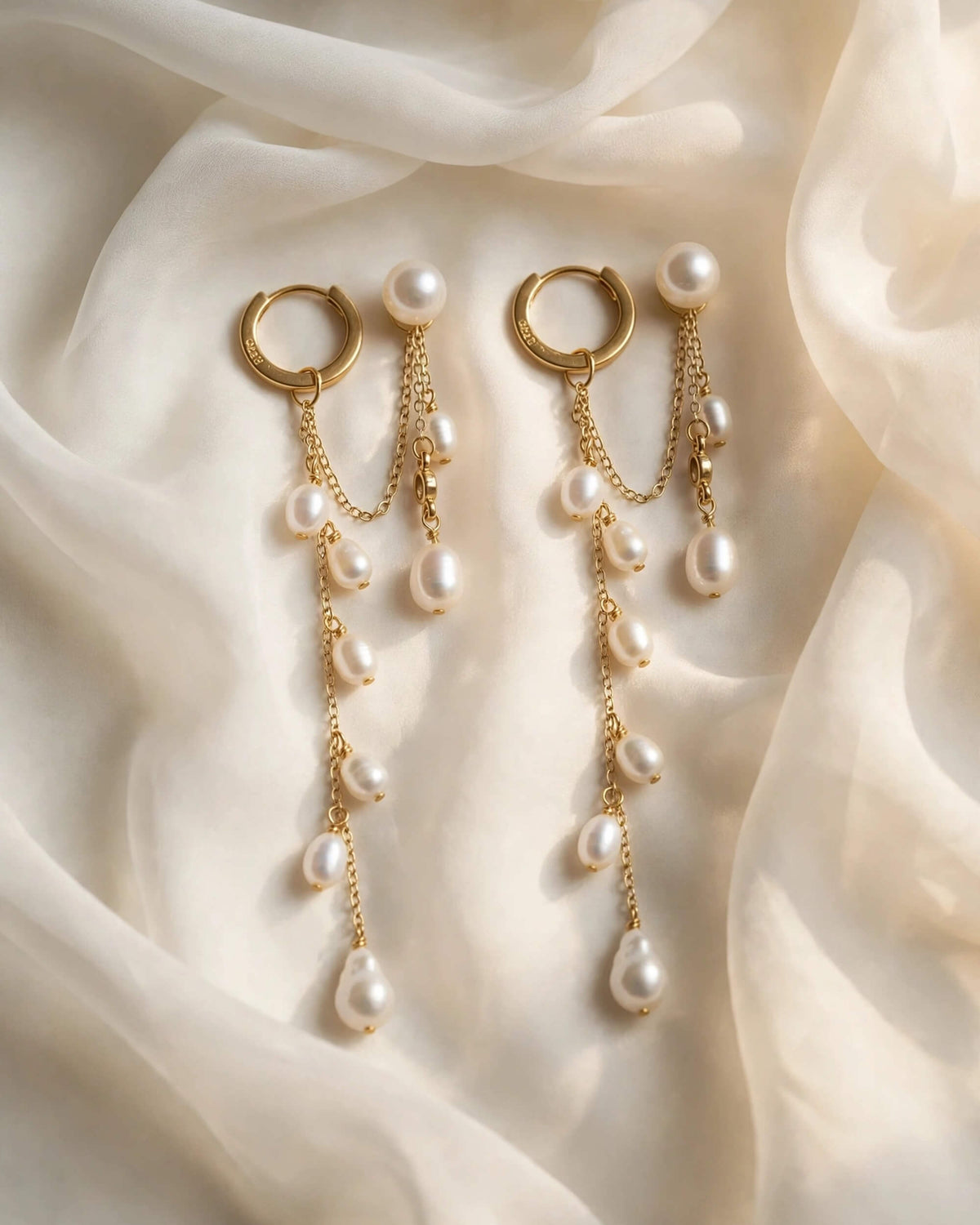 A pair of gold huggie hoop earrings with dangling chains and white freshwater pearls, styled on draped cream-colored silk fabric.