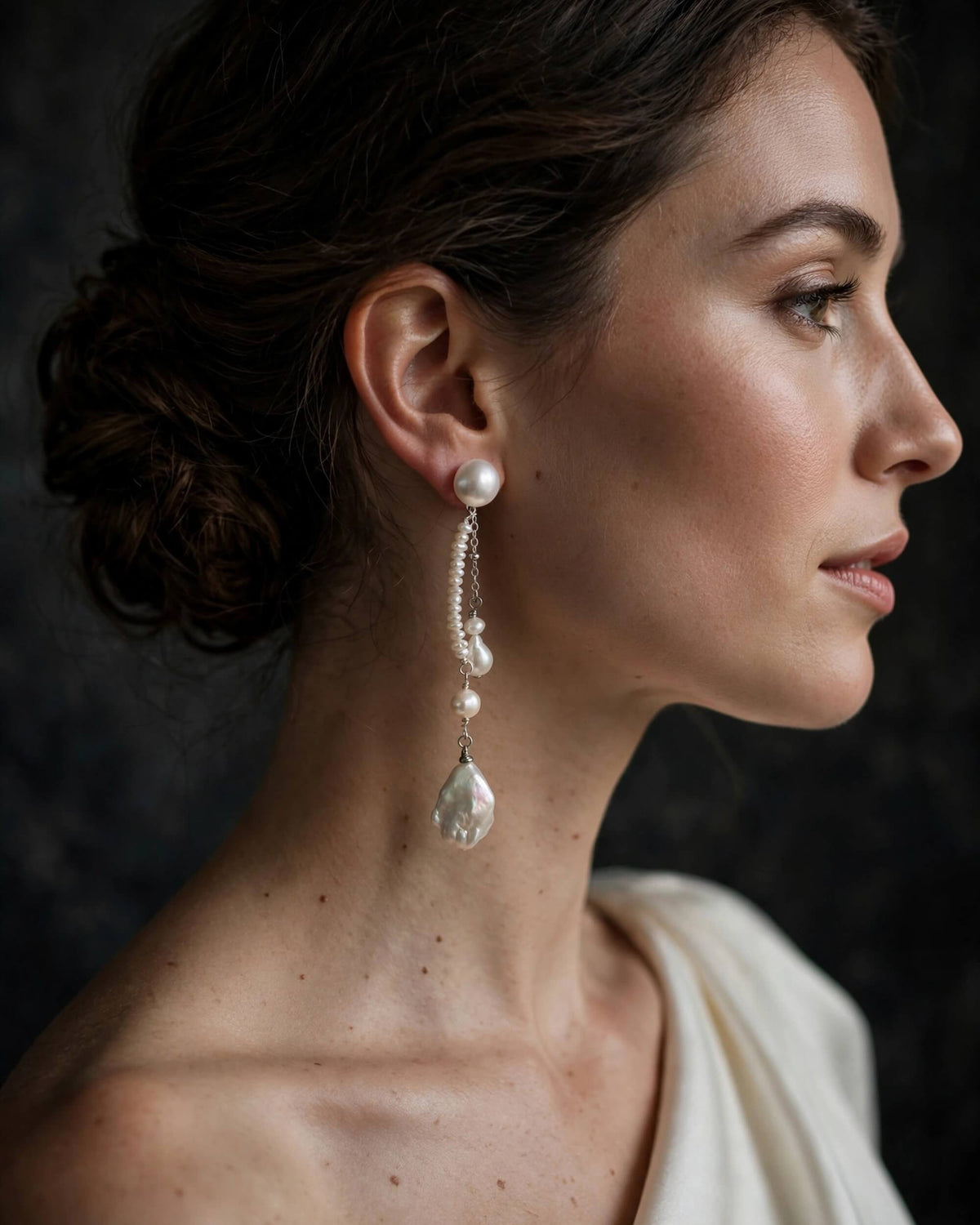 A close-up side profile of a woman wearing a long pearl drop earring with silver-toned chain links and a teardrop-shaped clear stone pendant, against a dark textured background.