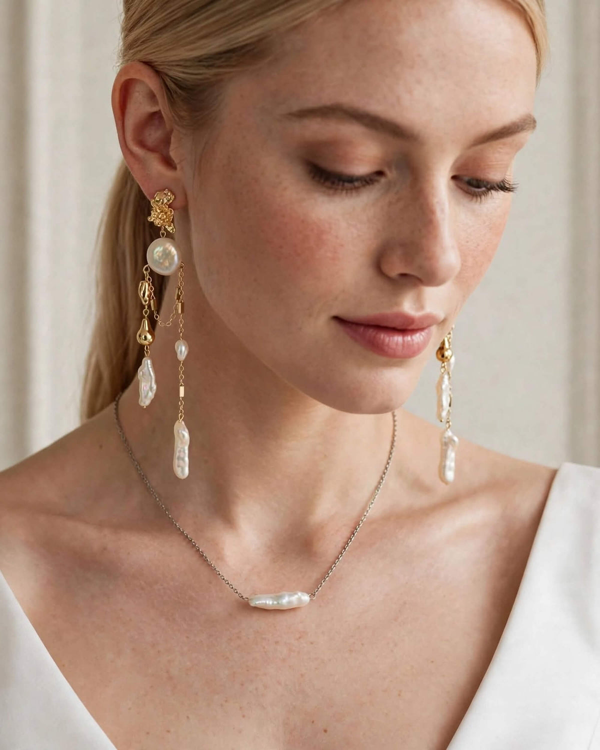 A close-up of a woman wearing elegant gold and pearl jewelry, including a baroque pearl necklace, matching drop earrings, and a delicate chain necklace, set against a soft beige background.