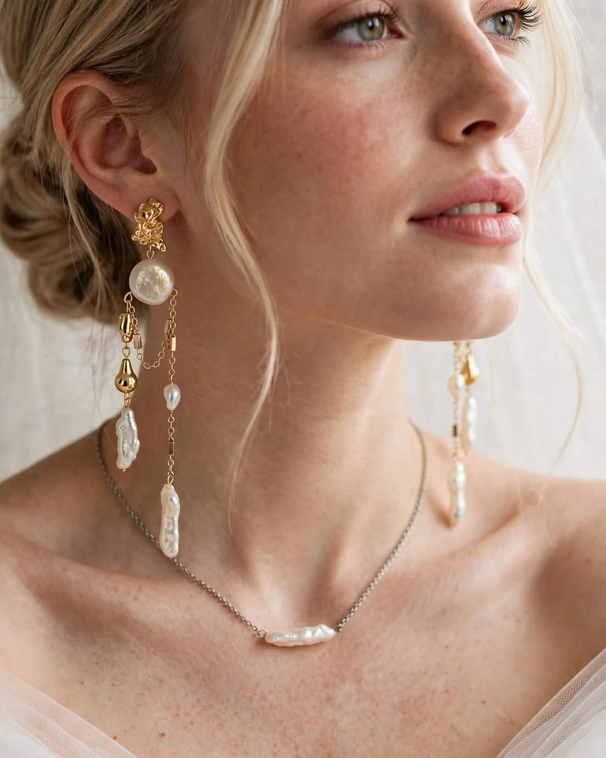 A close-up of a woman wearing elegant bridal jewelry, featuring a delicate silver chain necklace with a baroque pearl pendant and matching gold and pearl drop earrings.