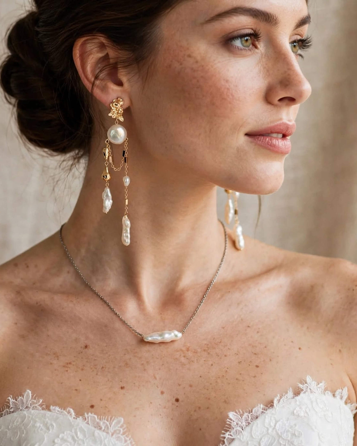 A close-up of a woman wearing bridal jewelry, including a silver chain necklace with a baroque pearl pendant and matching long gold and pearl drop earrings, paired with a white lace strapless top against a soft beige background.