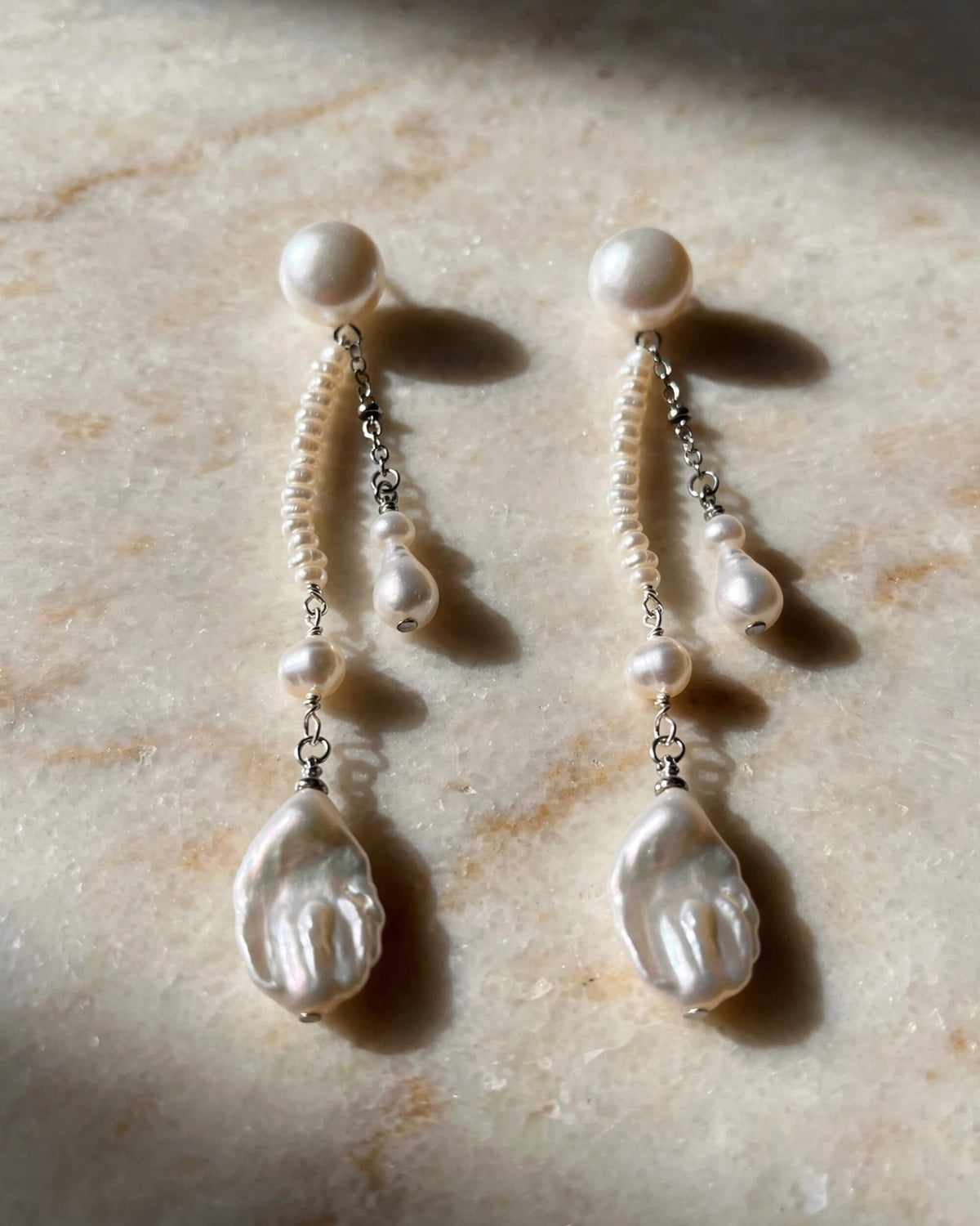 A pair of dangle earrings with baroque pearls and small white beads on silver chains, displayed on a light beige marble surface with soft shadows.