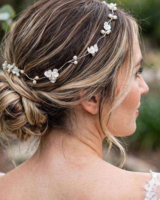 A woman in profile wearing a delicate silver wire headpiece adorned with small white floral charms and pearl-like beads, styled in an updo.