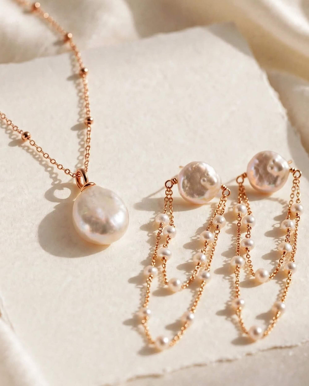 Rose gold jewelry set featuring a necklace and matching earrings with irregular baroque pearls and delicate chain details, displayed on a textured cream surface.