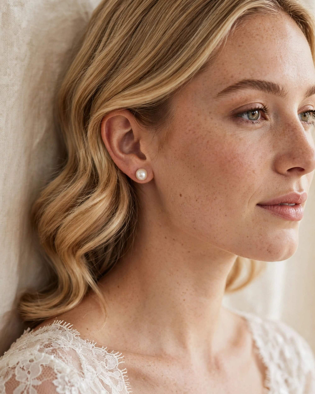 A close-up side profile of a woman with blonde hair wearing a small round pearl stud earring, leaning against a soft cream fabric background.