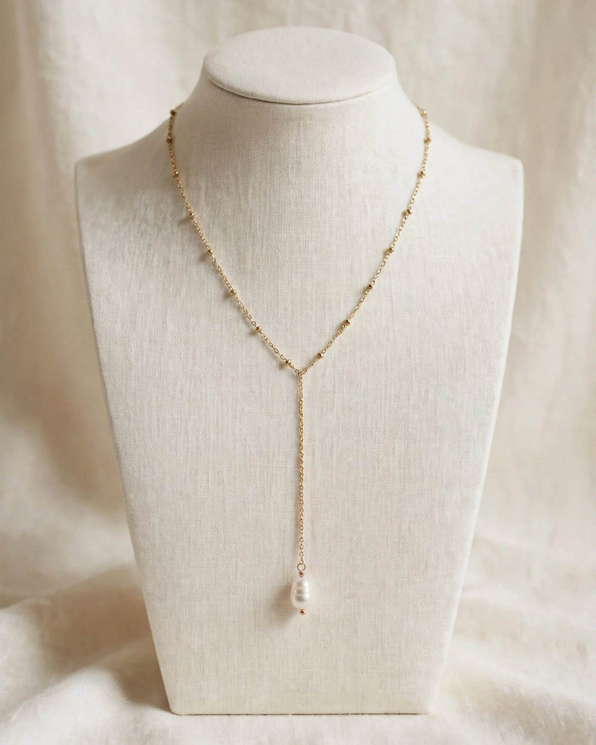 A delicate gold Y-necklace with small bead details and a single white pearl pendant, displayed on a cream-colored fabric jewelry bust.