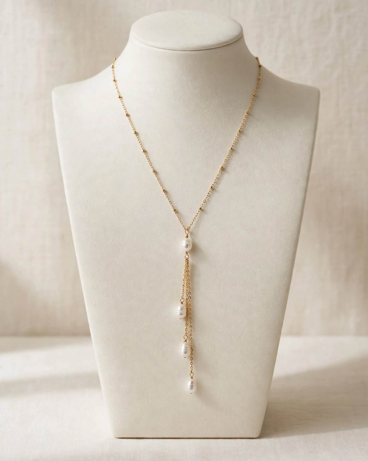 A delicate gold chain necklace with white pearl drops, displayed on a cream-colored jewelry bust against a soft beige background.
