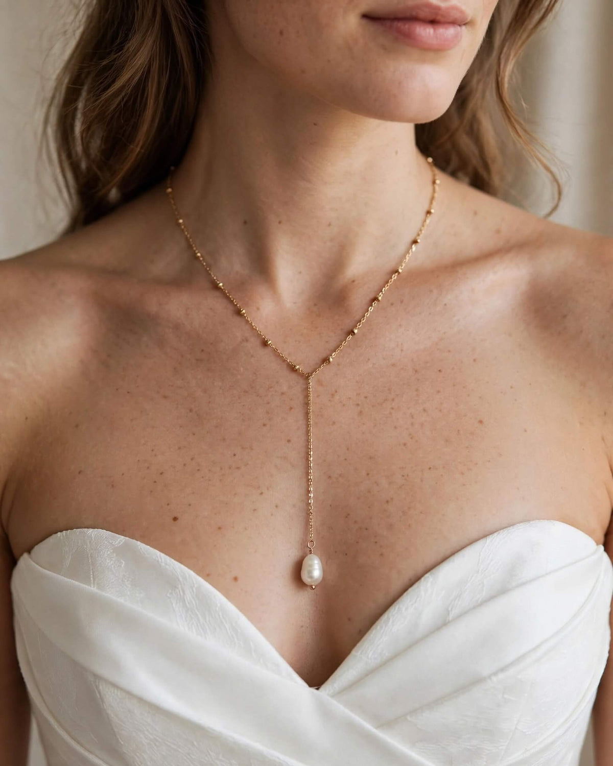 A close-up of a woman wearing a delicate gold lariat necklace with a single white pearl pendant, paired with a white strapless dress.
