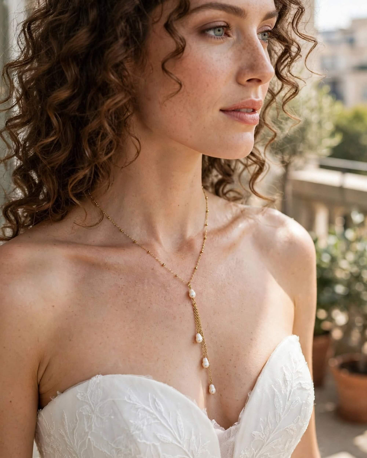 A woman with curly brown hair wearing a delicate gold lariat-style necklace with white pearl drops, paired with a white strapless lace dress in an outdoor setting.